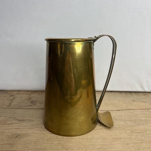 Könnte beinhalten: Ein antiker Messingbecher mit Henkel und Ausguss. Der Becher hat eine glänzende, polierte Oberfläche.