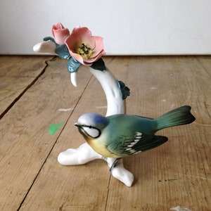 May include: A ceramic figurine of a bird perched on a white branch with pink flowers and green leaves. The bird has blue, green, and yellow coloring. The figurine is set against a wooden background.