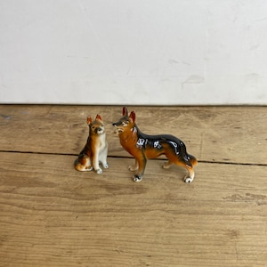 May include: Two ceramic German Shepherd dog figurines. One is sitting, the other is standing. The dogs are brown, black, and white with glossy finishes. The figurines are displayed on a wooden surface.