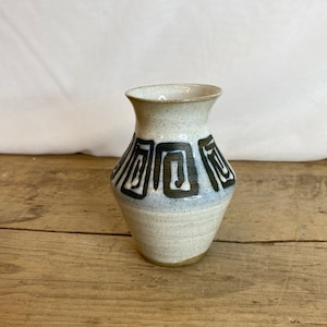 May include: A small white ceramic vase with a geometric black and brown design. The vase has a speckled glaze and a rounded base.