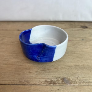 Puede incluir: Un pequeño cuenco de cerámica hecho a mano con un diseño único. El cuenco está dividido en dos secciones: una mitad es azul vibrante y la otra blanca. El cuenco se encuentra sobre una superficie de madera rústica, mostrando su naturaleza artesanal.