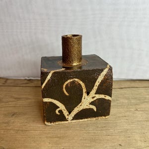 Pode incluir: Um castiçal quadrado de cerâmica com base castanha escura e um recipiente para velas dourado texturizado. O castiçal apresenta um design em espiral creme nas laterais. O recipiente para velas tem cerca de 5 cm de altura.