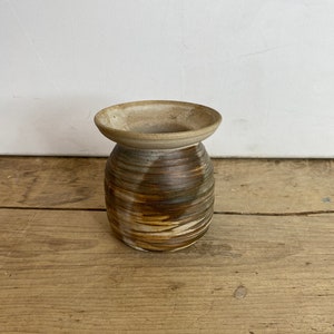 May include: A handmade ceramic vase with a wide, flared rim and a cylindrical body. The vase features horizontal bands of brown, tan, and white, creating a textured, earthy appearance. The vase is approximately 10 cm tall.