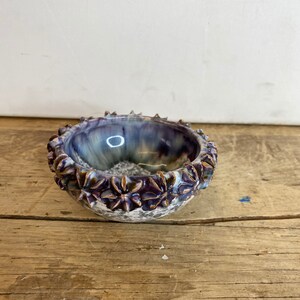 May include: A small, round ceramic bowl with a white, purple, and blue glaze. The bowl has a decorative rim with raised flower-shaped details.