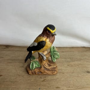 Puede incluir: Figurita de cerámica de un pájaro posado en una rama de árbol. El pájaro tiene la cabeza negra con una raya amarilla, cuerpo marrón y alas amarillas y negras. La rama es marrón con hojas verdes.