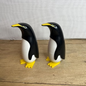 Könnte beinhalten: Zwei Pinguin-Figuren. Jeder Pinguin hat einen schwarzen Kopf und Rücken, einen weißen Bauch und gelbe Füße und einen Schnabel. Die Pinguine stehen auf einer Holzoberfläche.