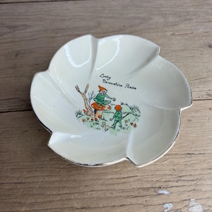 Könnte beinhalten: Cremefarbene, blütenblattförmige Schale mit Goldrand. Die Mitte zeigt eine skurrile Illustration von zwei Pixies mit dem Text "Lucky Devonshire Pixies". Die Schale steht auf einer Holzoberfläche.