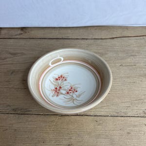 Puede incluir: Un cuenco de cerámica pequeño y redondo con exterior beige e interior blanco. El interior presenta un diseño floral con bayas rojas y tallos marrones. El cuenco tiene un asa pequeña y un anillo de acento rosa.