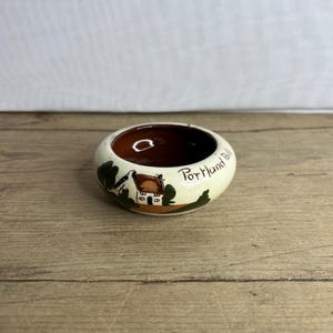 Op de afbeelding: Een kleine, ronde keramische kom met een bruin interieur en een crèmekleurige buitenkant. De buitenkant heeft een geschilderde scène van een huis en bomen, met de tekst "Portland Bill".
