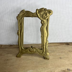 Op de afbeelding: Een sierlijke messingkleurige fotolijst met een Art Nouveau-ontwerp. De lijst heeft een rechthoekige opening en is versierd met bloemen en vrouwelijke figuren. De lijst staat op een houten oppervlak.