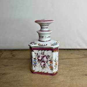 Puede incluir: Una botella de porcelana blanca con un diseño floral rosa y dorado. La botella tiene una base cuadrada y una parte superior redonda con un borde dorado.