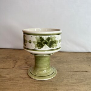 May include: A ceramic pedestal bowl with a cream interior and exterior. The bowl has a green floral design around the rim and on the exterior. The pedestal base is light green with darker green accents. The bowl is on a wooden surface.
