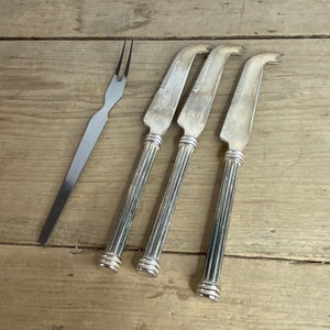 May include: A cheese knife set and a cheese fork. The knives have serrated blades and decorative handles. The fork and knives are silver-coloured and arranged on a wooden surface.