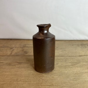May include: A small, brown ceramic bottle with a rounded body and a narrow, flared neck. The bottle has a dark, glossy finish and sits on a wooden surface. The bottle is likely an antique or vintage item.