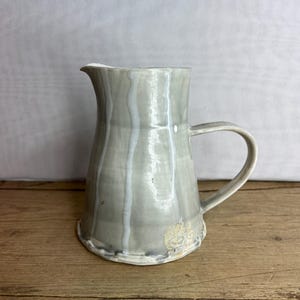 Puede incluir: Jarra de cerámica hecha a mano con un esmalte gris y vetas blancas. La jarra tiene un asa curva y un pico para verter. La base tiene un borde texturizado e irregular. La jarra está sobre una superficie de madera.