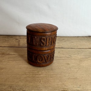 May include: A wooden spice jar with a lid. The jar is cylindrical with carved text that reads "ALL SPICE" and "CLOVES". The wood has a rich, dark brown color. The jar is sitting on a wooden surface.
