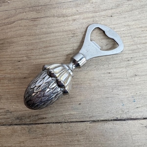 May include: A silver bottle opener with a decorative, artichoke-shaped handle. The opener is made of metal and has a classic design. The handle is detailed with textured patterns. The opener is designed for removing bottle caps.