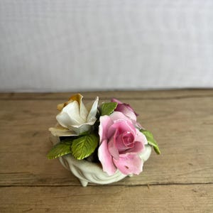 Może przedstawiać: Ceramiczna kompozycja kwiatowa z różowymi i białymi różami oraz zielonymi liśćmi, prezentowana w małej, kremowej miseczce z nóżkami. Kompozycja stoi na drewnianej powierzchni. Jest to przedmiot dekoracyjny.