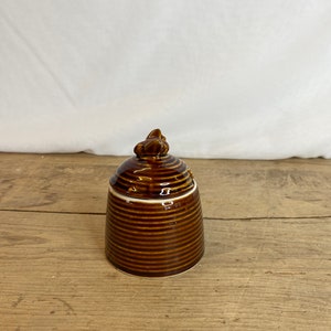 Könnte beinhalten: Ein brauner Keramiktopf mit Deckel. Der Topf hat ein geripptes Design und ein kleines dekoratives Element auf dem Deckel. Der Topf steht auf einer Holzoberfläche und einem weißen Hintergrund.