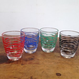 May include: Four clear shot glasses with colorful, wavy designs. Each glass features a different color: red, blue, green, and black. The glasses are arranged on a wooden surface.