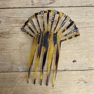 May include: A vintage hair comb with a fan-shaped design. The comb features a tortoiseshell pattern with clear rhinestone accents. The comb's teeth are long and slender, and the overall design is elegant and decorative.