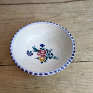 May include: Small, off-white ceramic bowl with a floral design. The bowl has a light blue rim with purple dots. The floral design includes red, yellow, and blue flowers with green leaves. The bowl sits on a wooden surface.