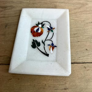 May include: A white marble rectangular dish with a floral inlay design. The inlay features a red and orange flower, green leaves, and blue and white accents. The dish has a slightly raised edge.