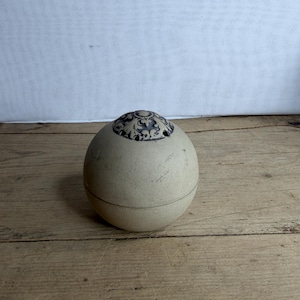 May include: A spherical ceramic container with a matte beige finish and a horizontal seam around the center. The top features a circular dark blue and white inlay with an intricate floral pattern and small ventilation holes along the edge.