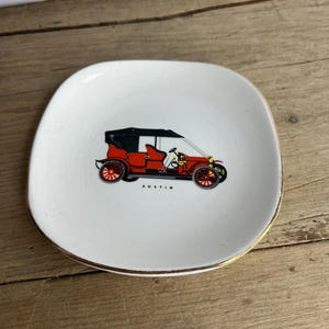 May include: A square, white ceramic dish with a gold rim. The dish features a red and black vintage car illustration with the word "AUSTIN" below the car. The dish is on a wooden surface.