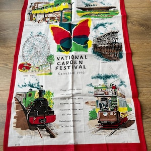 May include: A white tea towel with a red border. It features illustrations of a Ferris wheel, train, tram, and other attractions. The text "NATIONAL GARDEN FESTIVAL Gateshead 1990" is displayed, along with additional text and the words "MADE IN UK".