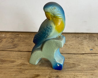 May include: A blue and yellow ceramic parrot figurine perched on a blue and white ceramic stand. The parrot is facing to the left and has a yellow beak.