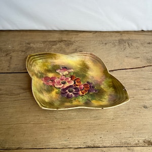 Puede incluir: Un plato antiguo en forma de hoja con un diseño floral pintado. El plato presenta un borde dorado y una imagen central de flores rosas, moradas y naranjas sobre un fondo verde y amarillo. El plato está sobre una superficie de madera.