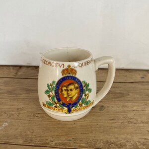 Może przedstawiać: Kremowy ceramiczny kubek z królewskim herbem z napisem "GEORGE VI QUEEN ELIZABETH" i mottem "DIEU ET MON-DROIT". Kubek ma uchwyt i lekko rozszerzony brzeg, z koroną i portretami.