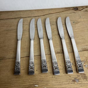 May include: Six silver dinner knives with ornate handles. The knives have a simple, straight blade and a decorative, embossed handle with a floral design.
