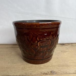 Könnte beinhalten: Ein brauner Keramiktopf mit Blumenmuster. Der Topf hat eine strukturierte Oberfläche und einen leicht ausgestellten Rand.