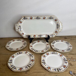 Puede incluir: Una fuente de porcelana blanca vintage y seis platos a juego. El conjunto presenta un diseño de borde floral y geométrico en naranja, morado y azul, con ribete dorado. La fuente es rectangular con asas y los platos son ovalados.
