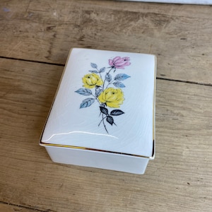 May include: A rectangular white ceramic trinket box with a floral design. The lid features yellow and pink roses with blue leaves. The box has gold trim and sits on a wooden surface.