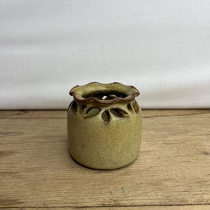 Peut inclure: Petit vase rond en céramique, vert clair et moucheté. Le vase présente un bord festonné et des découpes décoratives en forme de feuilles sur la partie supérieure. L'intérieur est brun foncé. Le vase est posé sur une surface en bois.