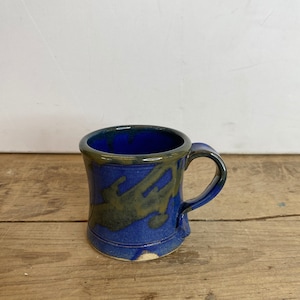 Peut inclure: Une tasse en céramique faite à la main, avec un glaçage bleu foncé et un motif vert olive contrastant. La tasse a un corps arrondi, une anse robuste et un bord légèrement évasé. Elle est posée sur une surface en bois.