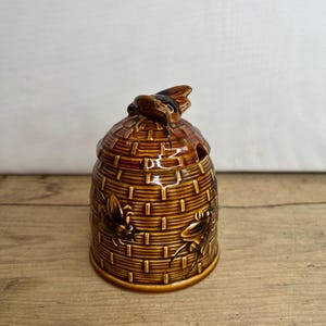 Puede incluir: Un tarro de miel de cerámica marrón con forma de colmena con una abeja en la parte superior. El tarro tiene una superficie texturizada y dos abejas en los lados.