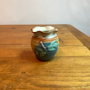 Puede incluir: Un pequeño jarrón de cerámica hecho a mano con un borde con volantes. El jarrón presenta una paleta de colores de color canela, azul y verde, con un diseño de hojas. El jarrón está sobre una superficie de madera.