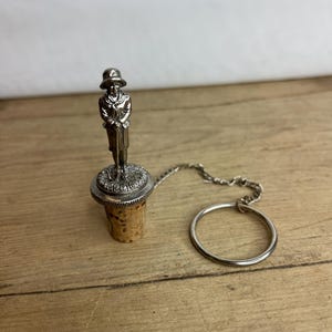 May include: Silver bottle stopper with a small figurine of a woman wearing a hat and a coat. The stopper is attached to a small silver chain with a ring.