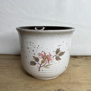 Puede incluir: Una maceta de cerámica con exterior blanco moteado e interior marrón oscuro. Un diseño floral rosa con hojas verdes decora el frente. La maceta tiene un borde ligeramente ensanchado y está sobre una superficie de madera.