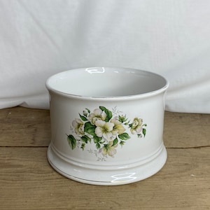 May include: White ceramic planter with a floral design of white and yellow flowers and green leaves. The planter has a wide, cylindrical shape with a flared base and gold trim. Measures approximately 15 cm in diameter.