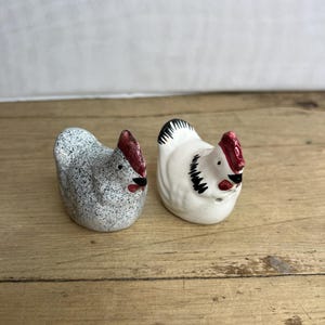 May include: Two ceramic chicken figurines. One is speckled gray, the other is white. Both have red combs and beaks, and are set on a wooden surface. These decorative figurines are likely for kitchen or home decor.