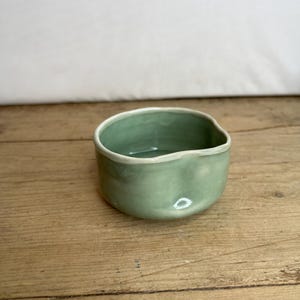 May include: A small, green ceramic bowl with a slightly irregular rim. The bowl is sitting on a wooden surface.