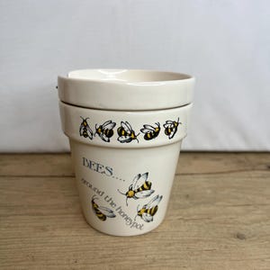 Könnte beinhalten: Ein cremefarbener Honigtopf aus Keramik mit Deckel, verziert mit schwarz-gelben Bienenillustrationen. Der Topf trägt den Text "BEES... around the honeypot" in Blau und Schwarz. Der Topf steht auf einer Holzoberfläche.