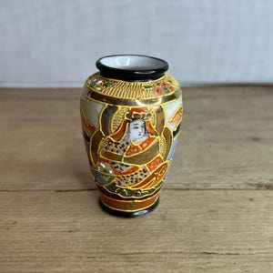 以下が含まれることがあります： 黒い縁と底を持つ、小さな装飾的な日本の花瓶。伝統的な衣装を着た人物の精巧なデザインが特徴で、金色のアクセントとカラフルな模様が施されています。背景はニュートラルカラーです。
