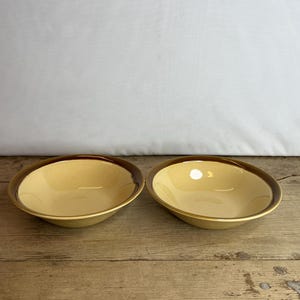 Puede incluir: Dos cuencos de cerámica con interior amarillo claro y borde marrón oscuro. Los cuencos están colocados sobre una superficie de madera. Los cuencos están vacíos y listos para ser llenados con comida.