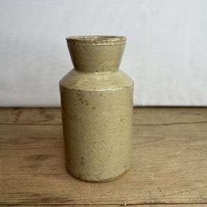 Puede incluir: Un jarrón de gres beige con cuerpo cilíndrico y cuello acampanado. El jarrón tiene una superficie texturizada y un esmalte de color claro. Este jarrón vintage es adecuado para la decoración de la cocina o del hogar.